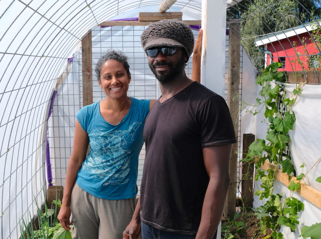Deepa and Victor stand in a small greenhouse they built