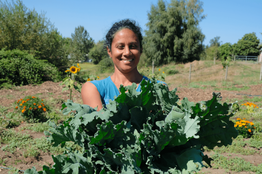 Deepa smiles and holds out kale on a sunny day at Keerai Farm