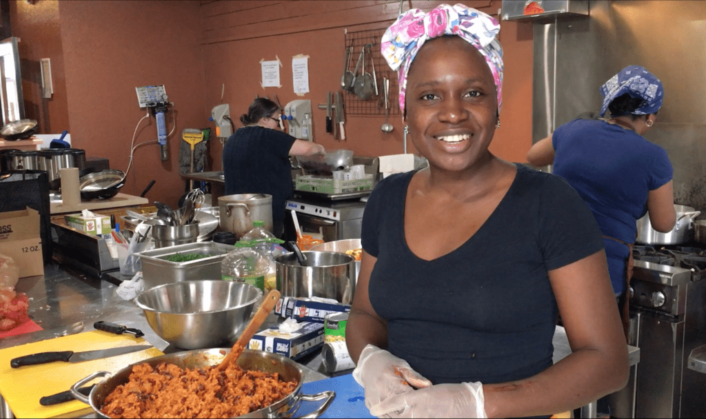 Lilian Ryland smiles at the camera. She stands in a busy kitchen next to a big pot of beans and rice.